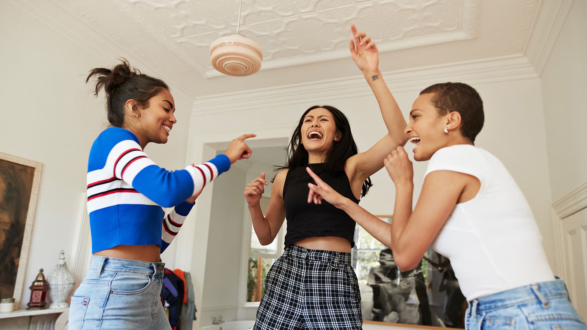 Three women enjoying a lively moment indoors, dancing and laughing with casual attire and an elegant ceiling design overhead.