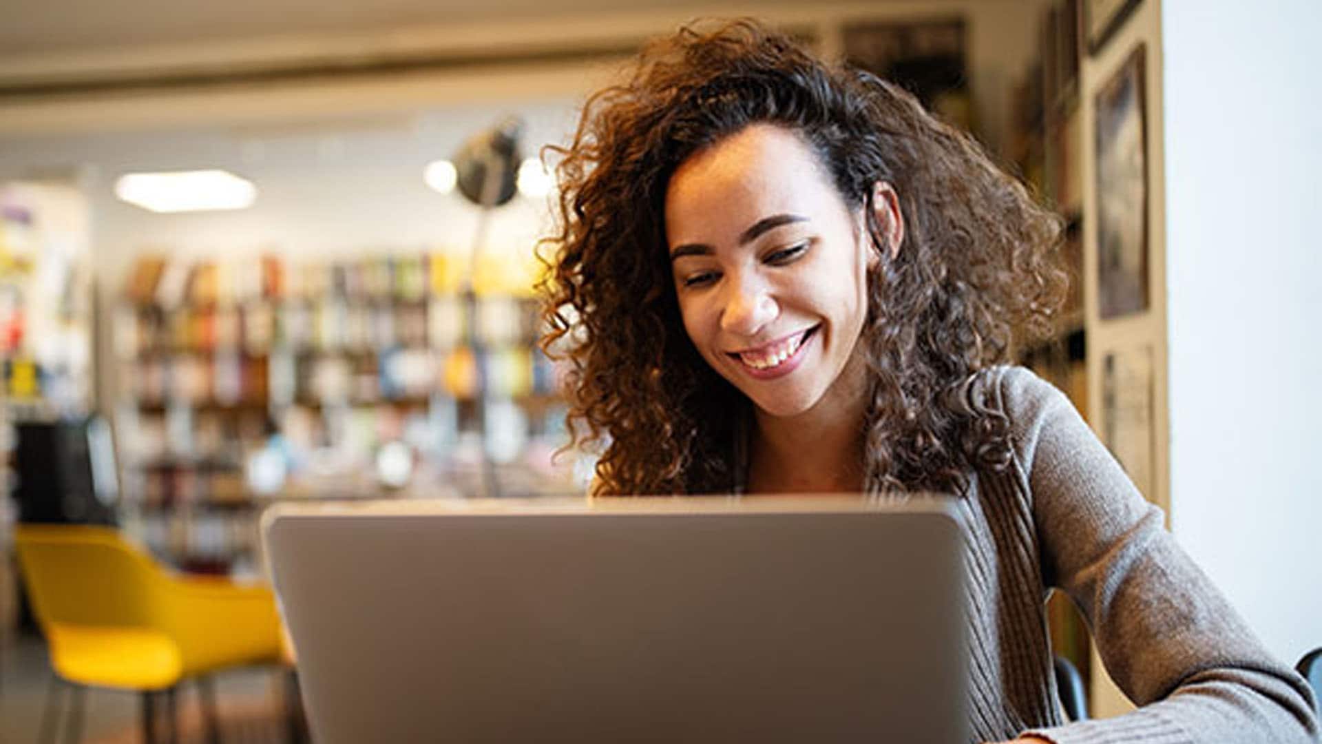 A woman sitting in a chair, smiling at a laptop on a table, with a wall and furniture visible in the background.