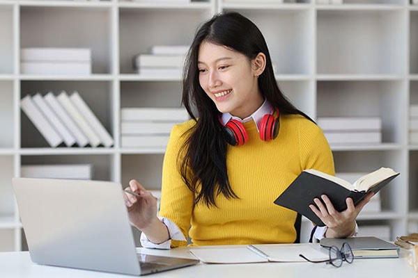 A woman in a yellow sweater with red headphones smiles while using a laptop and holding a book. She's seated at a desk with shelves in the background.